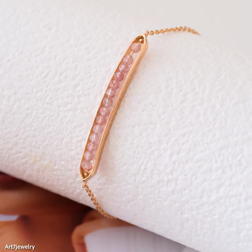 Gold bracelet with pink beads on a white surface, brand 'Art7Jewelry' visible.