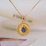 Gold sapphire pendant necklace with natural blue sapphire center stone, surrounded by small sparkling accents on a textured round gold disc, delicate minimalist chain jewelry for women