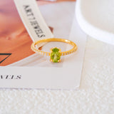 Oval green peridot solitaire ring in a gold-tone four-prong setting with sparkling clear stone band accents.