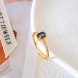 Blue sapphire solitaire ring with an oval deep-blue center stone, gold-tone four-prong setting, and sparkling clear stone band accents.