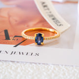 Blue sapphire solitaire ring with an oval deep-blue center stone, gold-tone four-prong setting, and sparkling clear stone band accents.