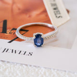 Blue sapphire solitaire ring with an oval deep-blue center stone, silver-tone four-prong setting, and a slim clear stone accent band.