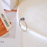 Blue sapphire solitaire ring with an oval deep-blue center stone, silver-tone four-prong setting, and a slim clear stone accent band.