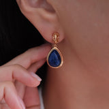 A close-up of a gold teardrop dangle earring with a deep blue gemstone, worn on a person's ear with a hand adjusting it nearby.